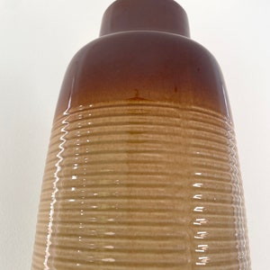 May include: A tall, brown ceramic vase with a ribbed texture. The top portion is a darker shade of brown, transitioning to a lighter tan color on the lower, textured body. The vase has a glossy finish.