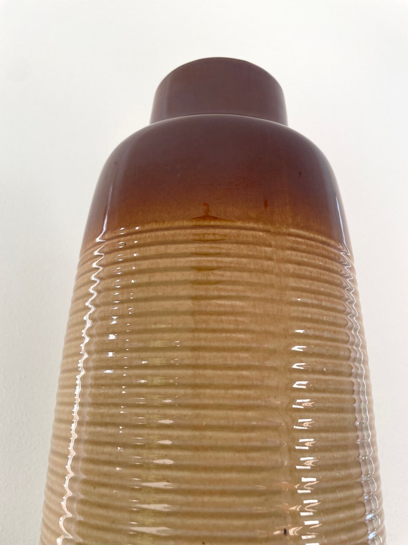 May include: A tall, brown ceramic vase with a ribbed texture. The top portion is a darker shade of brown, transitioning to a lighter tan color on the lower, textured body. The vase has a glossy finish.