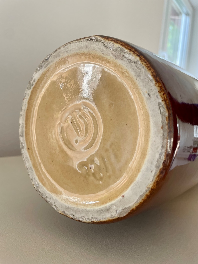 May include: A brown ceramic jug with a glossy finish. The base is light tan with a white rim and a stamped design. The jug's exterior is a deep brown color.