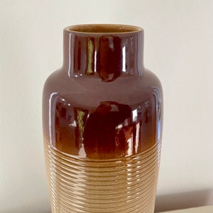 May include: A tall, brown ceramic vase with a glossy finish. The upper portion is a darker shade of brown, transitioning to a lighter, textured tan base with horizontal ridges. The vase has a wide, open top.