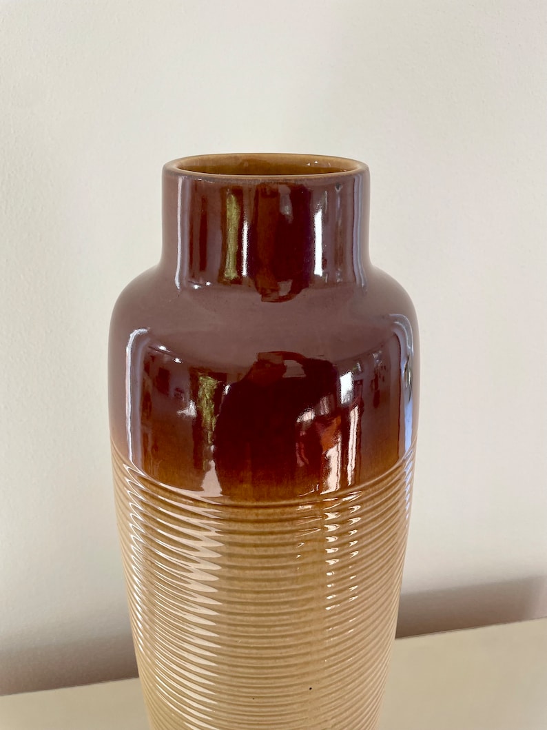 May include: A tall, brown ceramic vase with a glossy finish. The upper portion is a darker shade of brown, transitioning to a lighter, textured tan base with horizontal ridges. The vase has a wide, open top.
