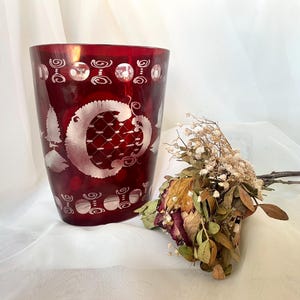 May include: A ruby red glass vase with etched white floral and geometric designs. The vase has a wide, cylindrical shape and is decorated with a large, stylized floral motif. A small bouquet of dried flowers rests beside the vase.