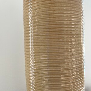 May include: A tall, cylindrical ceramic vase with a textured, ribbed design. The vase has a light beige body with a darker brown rim. The surface reflects light, highlighting the horizontal ridges.