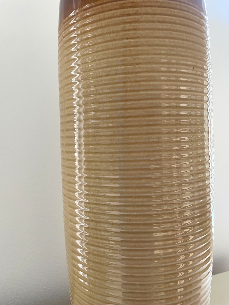 May include: A tall, cylindrical ceramic vase with a textured, ribbed design. The vase has a light beige body with a darker brown rim. The surface reflects light, highlighting the horizontal ridges.