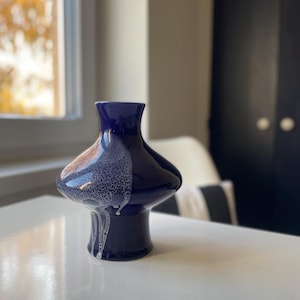 Può includere: Un vaso in ceramica blu scuro dalla forma unica. Il vaso ha una parte superiore ampia e arrotondata e una base più stretta. La glassa bianca maculata cola lungo il lato, creando un contrasto sorprendente. Il vaso è posto su una superficie bianca.