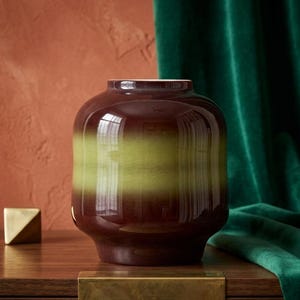 May include: A glossy, brown vase with a wide, cylindrical shape and a horizontal band of olive green. The vase sits on a wooden surface, with a gold-colored cube in the background. A green velvet curtain is in the background.