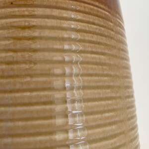 May include: Close-up of a tan and brown ceramic jug. The jug has a ribbed texture and a glossy finish. The top of the jug is a darker brown color.