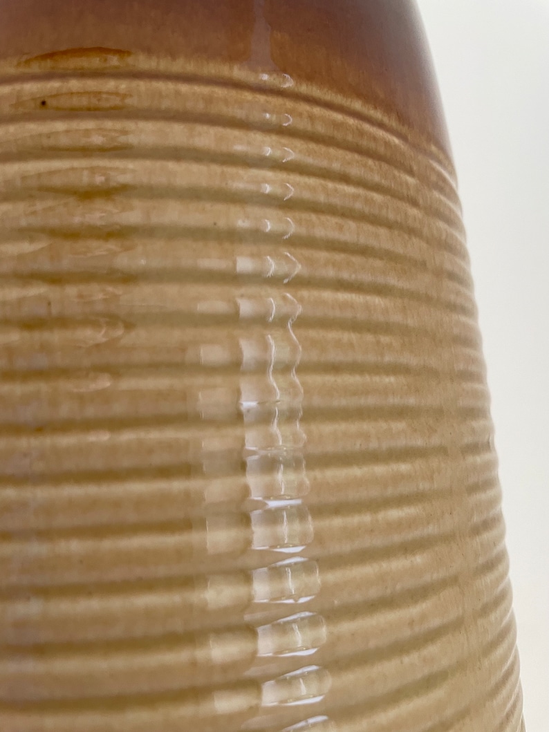 May include: Close-up of a tan and brown ceramic jug. The jug has a ribbed texture and a glossy finish. The top of the jug is a darker brown color.