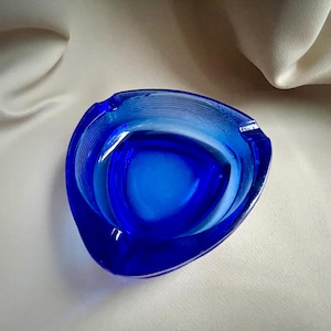 Puede incluir: Un cenicero azul vibrante con forma triangular. El cenicero es de vidrio y presenta un diseño estriado en el exterior. El interior tiene una depresión lisa y redondeada. Una pieza clásica para la decoración del hogar.