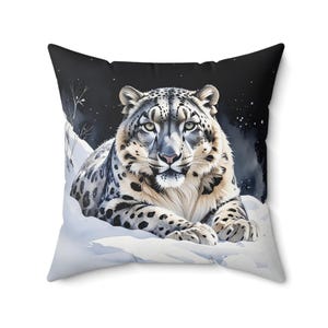 May include: Square throw pillow featuring a snow leopard illustration. The design showcases the big cat resting in a snowy landscape under a starry night sky. The pillow has a black background and a white border.