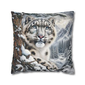 May include: Square throw pillow featuring a detailed illustration of a snow leopard. The big cat has white fur with black spots, blue eyes, and a pink nose. The background shows a snowy mountain landscape and evergreen trees.