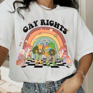 Puede incluir: Camiseta blanca con el texto "GAY RIGHTS" sobre un arcoíris. El gráfico presenta dos ranas tocando discos, rodeadas de flores, setas y notas musicales. La camiseta es de estilo casual y unisex.