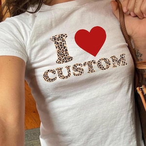 Può includere: T-shirt bianco con la scritta "I ♥ CUSTOM". La lettera "I" e la parola "CUSTOM" sono in un motivo leopardato. Un cuore rosso è tra la "I" e "CUSTOM". La maglietta è a girocollo.
