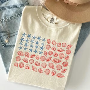 May include: A cream-colored t-shirt featuring a graphic of an American flag composed of blue starfish and red seashells. The shirt has a crew neckline and a Comfort Colors brand label printed on the inside of the collar area.