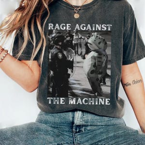 Può includere: T-shirt grigio scuro con la scritta "RAGE AGAINST THE MACHINE" sopra e sotto un'immagine in bianco e nero di una protesta. L'immagine presenta una persona in costume da rana e agenti di polizia.
