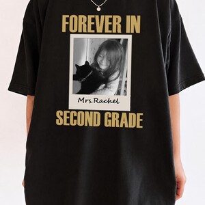 Może przedstawiać: Czarny t-shirt ze złotym napisem FOREVER IN na górze i SECOND GRADE na dole. W środku znajduje się kwadratowe czarno-białe zdjęcie dziecka trzymającego kota, oprawione jak polaroid z odręcznym napisem Mrs. Rachel.