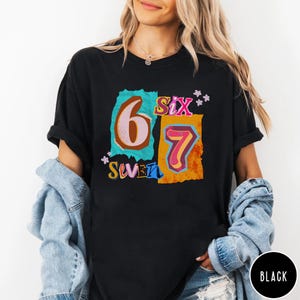 May include: Black t-shirt with a colorful graphic design featuring the numbers "6" and "7" in a retro style. The word "Six" and "Seven" are also included in the design. The shirt is a casual, everyday wear item.