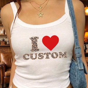 May include: White tank top with the text "I ❤️ CUSTOM". The letter "I" and the word "CUSTOM" are in a leopard print pattern. A red heart is between them. A gold necklace with star pendants is worn.