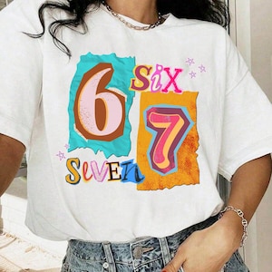 May include: White t-shirt with a colorful graphic design featuring the numbers "6" and "7" and the words "Six" and "Seven". The numbers are in a retro style with a mix of colors, including pink, blue, and orange. The shirt is worn with blue jeans.