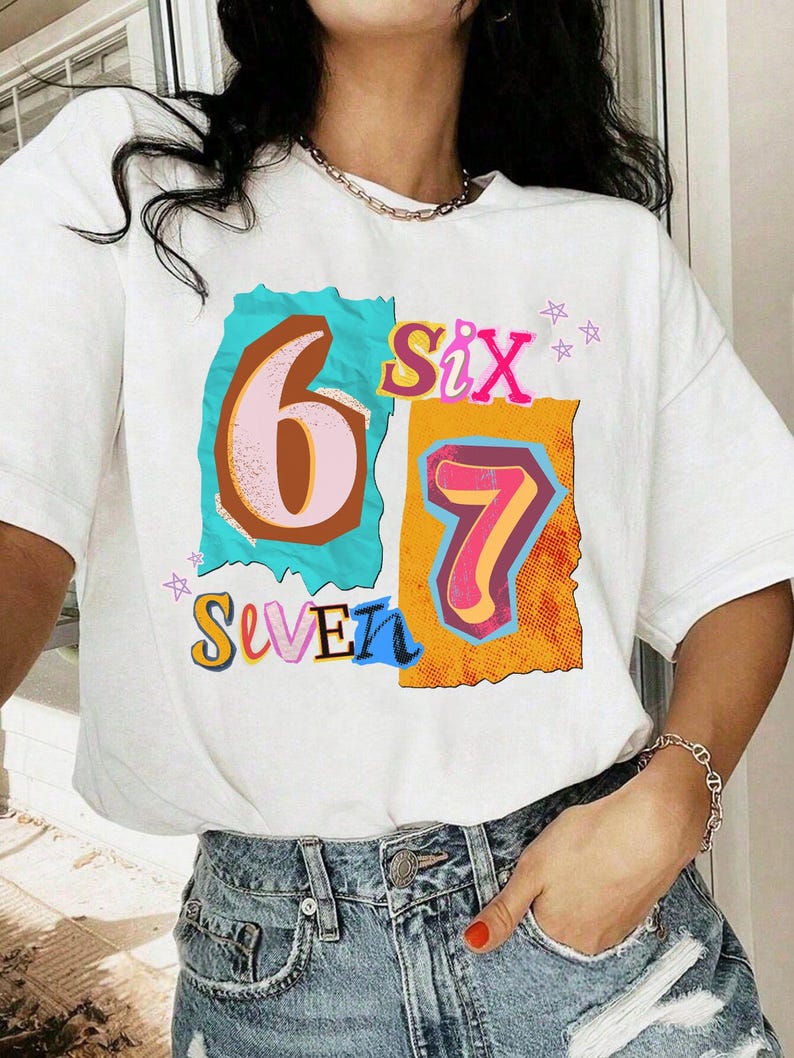 May include: White t-shirt with a colorful graphic design featuring the numbers "6" and "7" and the words "Six" and "Seven". The numbers are in a retro style with a mix of colors, including pink, blue, and orange. The shirt is worn with blue jeans.
