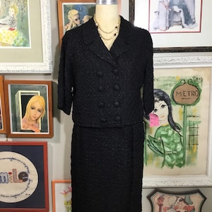 May include: A black textured skirt suit with a double-breasted jacket and a pearl necklace. The suit is on a mannequin in front of a wall with framed artwork.