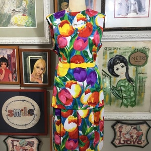 May include: A sleeveless dress with a vibrant floral tulip print in red, yellow, purple, and white on a blue background. The dress features a yellow belt and is displayed on a mannequin. A necklace with blue and white beads is also visible.