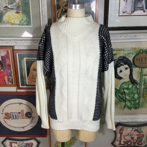 May include: White and black knit sweater with a mock neck and cable knit design. The sweater has a black and white geometric pattern on the sleeves.