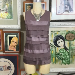 May include: A mauve sleeveless top with a layered ruffle design. The top has a satin trim and is displayed on a mannequin wearing a silver necklace.