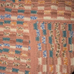 May include: A handmade crochet blanket in a patchwork design. The blanket features a repeating pattern of squares in shades of pink, blue, white, orange, and brown. The texture is visible, showcasing the individual stitches. The blanket is a cozy home decor item.