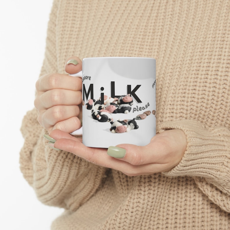 A woman holding an 11oz mug. The design on the mug is a group of cartoon cow shaped cookies lazily laying around with the phrase "moooore milk please" graphically designed to overlay on the photograph.