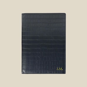 Op de afbeelding: Een rechthoekige donkerblauwe notitieboekhoes met een gestructureerd krokodillenleerpatroon. In de rechterbenedenhoek staan de initialen J.M. in een metallic gouden lettertype. De hoes heeft een strakke, gestructureerde rand en een plat oppervlak.