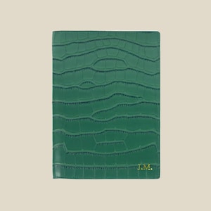 Può includere: Un taccuino in pelle verde con un motivo a coccodrillo in rilievo e le iniziali "J.M." incise in oro sulla copertina.