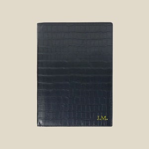 Puede incluir: Una carpeta de cuero negro con un patrón de cocodrilo en relieve y las iniciales "J.M." en letras doradas.