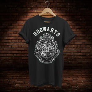 Puede incluir: Camiseta negra con un escudo blanco de Hogwarts que representa el escudo de la Escuela de Magia y Hechicería de Hogwarts. El escudo incluye un león, un águila, un tejón y una serpiente con el texto "Draco Dormiens Nunquam  Titillandus".