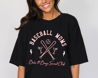 Op de afbeelding: Zwart oversized T-shirt met een wit grafisch ontwerp met de tekst "Baseball Moms Bats & Booze Social Club". De grafiek toont twee gekruiste honkbalknuppels, een honkbal en twee martini glazen.