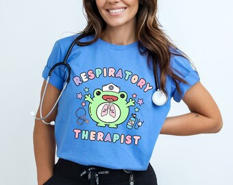 Pode incluir: Uma camiseta azul claro com um sapo de desenho animado usando um chapéu de enfermeira e um estetoscópio. O texto "Respiratory Therapist" está impresso na camiseta.