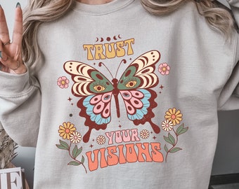 May include: A light gray sweatshirt with a retro-style butterfly graphic and the text "TRUST YOUR VISIONS". The butterfly has large eyes and is surrounded by flowers.