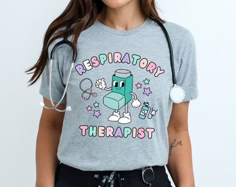 Pode incluir: Uma camiseta cinza com um inalador de desenho animado usando um estetoscópio e segurando uma garrafa de medicamento. O texto "Respiratory Therapist" está impresso na camiseta.