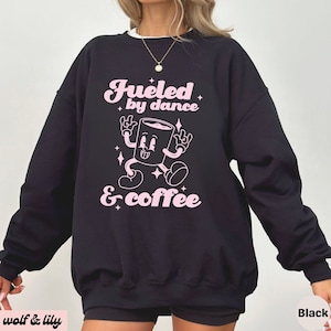 Peut inclure: Sweat-shirt noir avec l'inscription rose "Fueled by dance & coffee". Le motif représente un personnage de tasse à café de dessin animé. Le sweat-shirt a un col blanc et est de style décontracté et confortable.