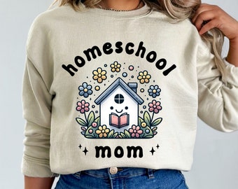 Puede incluir: Una sudadera beige con el texto "homeschool mom" en letras negras. El diseño presenta una casa de dibujos animados con una cara sonriente que sostiene un libro, rodeada de flores de colores.