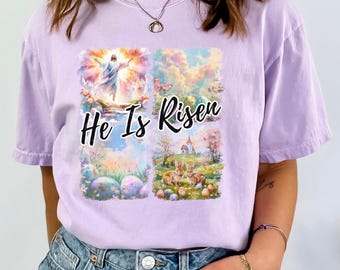 May include: A light purple t-shirt featuring a colorful graphic with the text "He Is Risen." The graphic includes images of Jesus, Easter eggs, butterflies, and bunnies, set against a sky background.