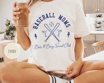 Op de afbeelding: Een wit T-shirt met de tekst "Baseball Moms" in blauwe letters. Het shirt heeft ook een honkbalknuppel en een bal, gekruist met een martini-glas aan elke kant. De tekst "Bats & Booze Social Club" staat onder de afbeelding gedrukt. Het shirt wordt getoond op een model dat het draagt.