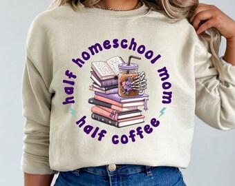 Puede incluir: Una sudadera beige con el texto "half homeschool mom half coffee" en letras moradas. El texto está rodeado por un círculo y una pila de libros con un tarro de cristal de café en la parte superior.