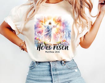 May include: A white t-shirt with a colorful image of Jesus Christ rising from the grave. The text "He is risen" and "Matthew 28:6" is printed below the image.