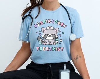 Pode incluir: Camiseta azul claro com um guaxinim de desenho animado usando um chapéu de enfermeira e equipamentos médicos. As palavras "RESPIRATORY THERAPIST" estão acima e abaixo do guaxinim. Um estetoscópio está pendurado no pescoço.