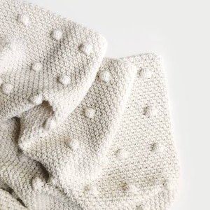 May include: A white crocheted throw blanket with a textured, bobble stitch pattern. The blanket is folded in half and has a soft, cosy look.