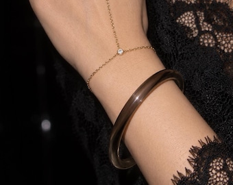 May include: A gold hand chain featuring two small round clear stones set in metal, connected by a delicate chain that links a finger ring to a wrist bracelet. A smooth, dark translucent bangle is worn on the wrist below the hand chain.