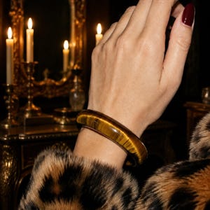 May include: A smooth, polished tiger's eye gemstone bangle bracelet featuring a rounded profile and a distinct chatoyant golden-brown banded pattern that reflects light across its curved, solid surface.