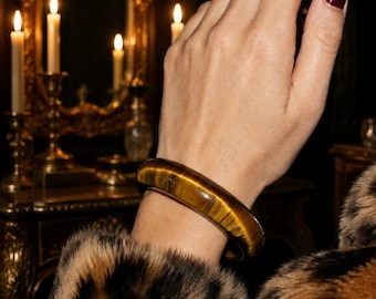 May include: A smooth, polished tiger's eye gemstone bangle bracelet featuring a rounded profile and a distinct chatoyant golden-brown banded pattern that reflects light across its curved, solid surface.