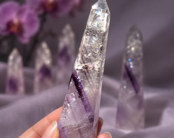 May include: A clear, faceted crystal tower featuring internal purple inclusions and fractures that catch the light. The stone has a pointed top and a rectangular base, displaying natural variations in transparency and deep violet color bands.